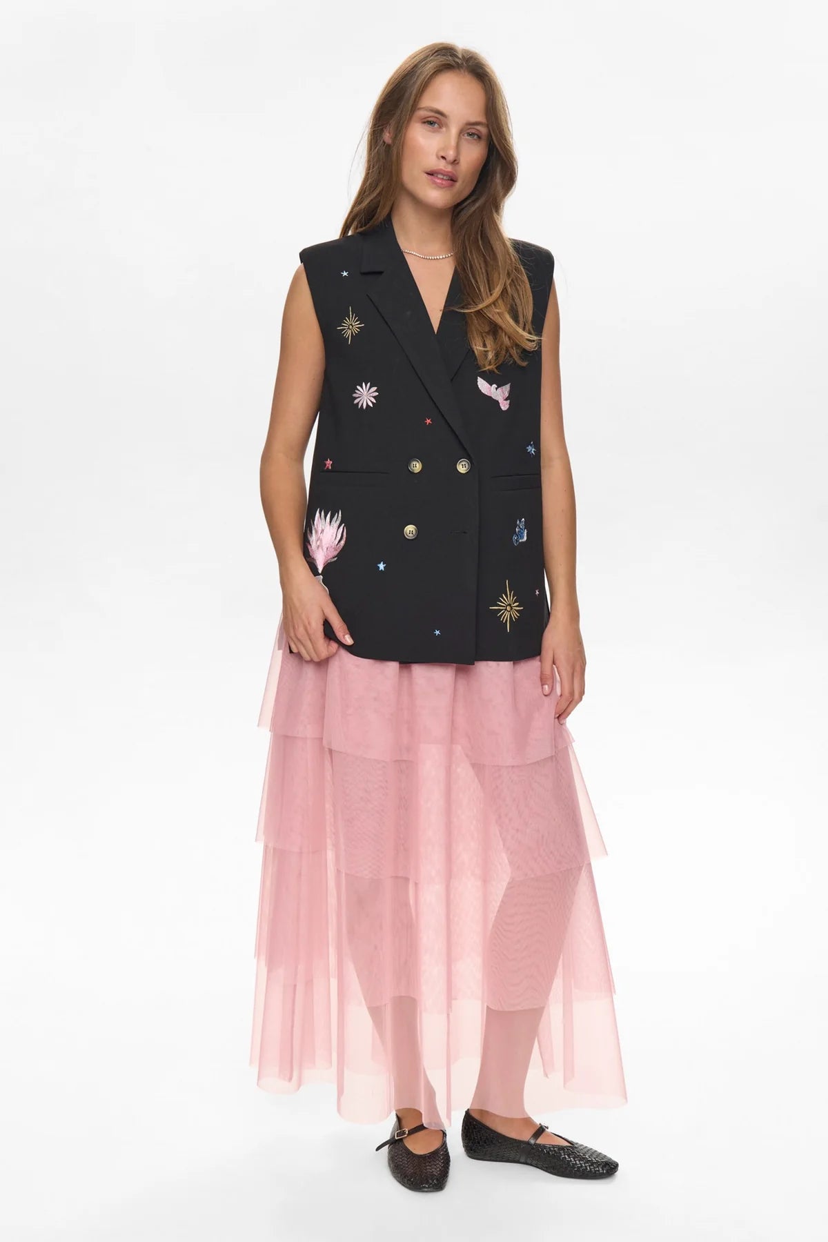 Woman wearing a black sleeveless jacket with floral embroidery and a pink skirt on a white background