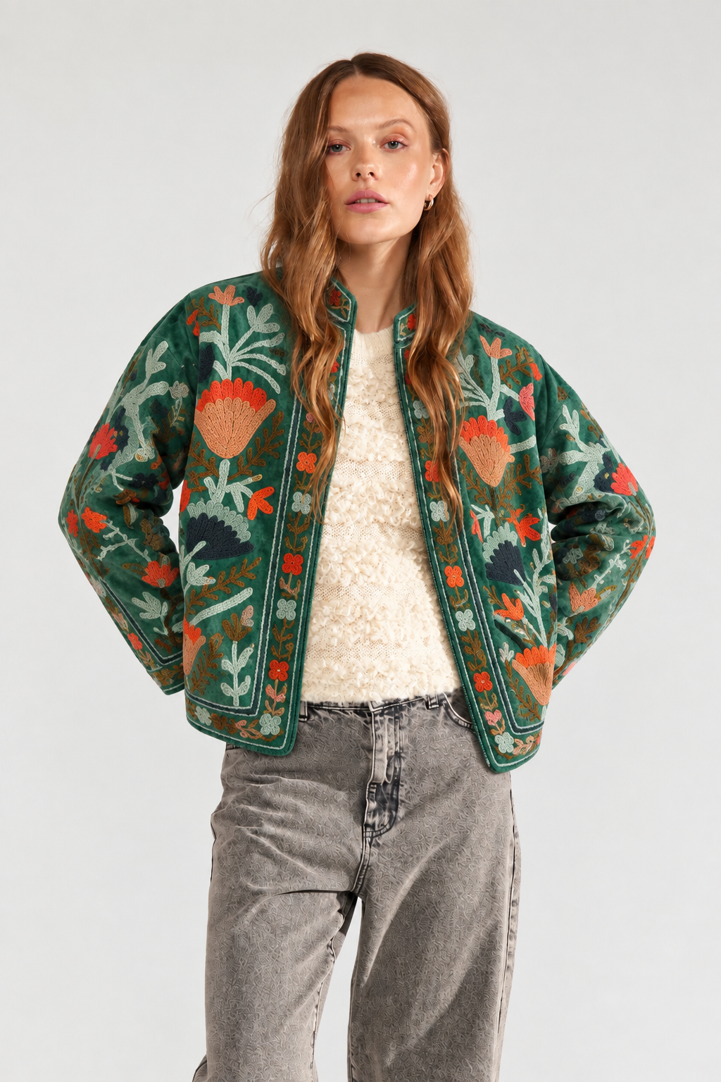 A woman modeling a green embroidered bomber jacket with orange and red floral motifs, worn over a cream textured top and gray jeans, against a light gray studio background.