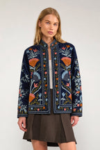 A woman modeling a navy blue embroidered jacket with colorful floral patterns, paired with a brown pleated skirt, against a plain light gray background.