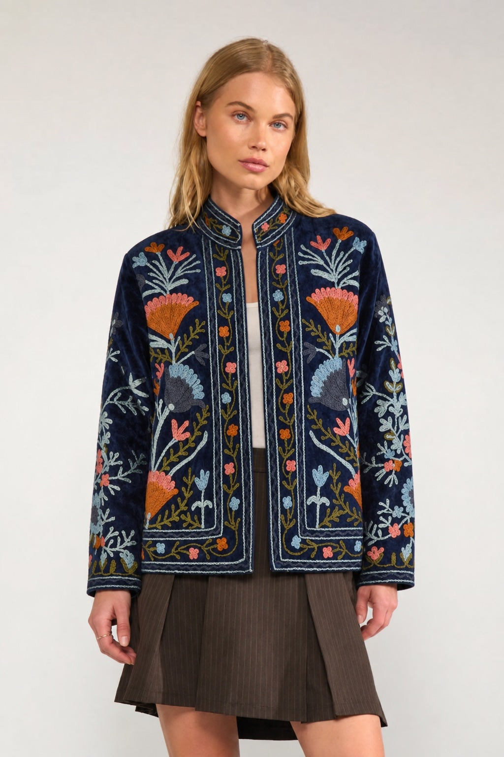 A woman modeling a navy blue embroidered jacket with colorful floral patterns, paired with a brown pleated skirt, against a plain light gray background.