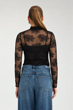 A woman viewed from the back wearing a black sheer floral lace long-sleeve top over a black camisole, paired with blue jeans, against a light gray studio background.
