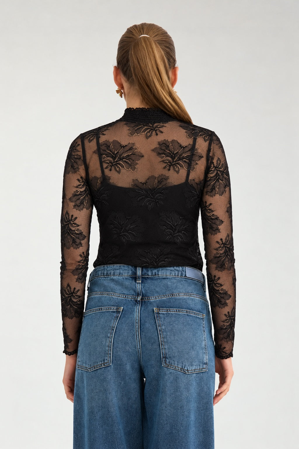 A woman viewed from the back wearing a black sheer floral lace long-sleeve top over a black camisole, paired with blue jeans, against a light gray studio background.
