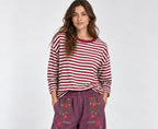 A woman modeling a red and white striped long-sleeve top with purple wide-leg pants featuring floral embroidery on the front pockets, paired with burgundy slip-on shoes, against a light gray studio background.