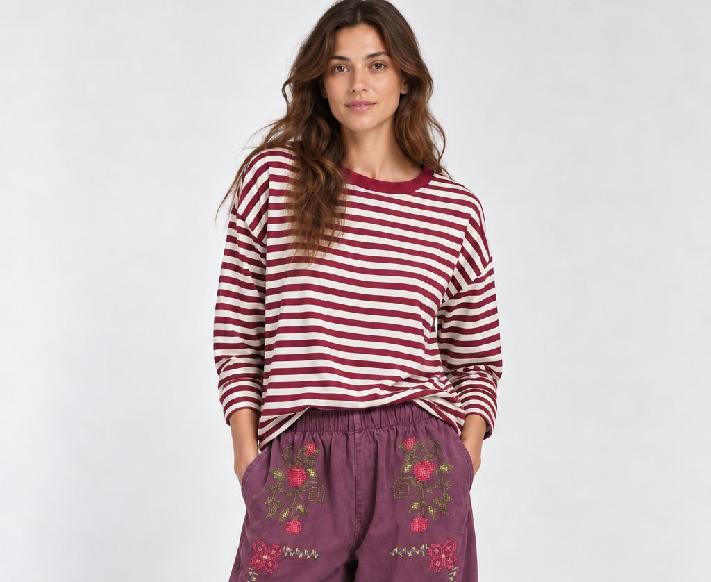 A woman modeling a red and white striped long-sleeve top with purple wide-leg pants featuring floral embroidery on the front pockets, paired with burgundy slip-on shoes, against a light gray studio background.
