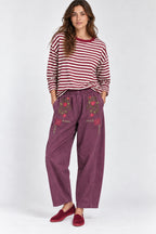 A woman modeling a red and white striped long-sleeve top with purple wide-leg pants featuring floral embroidery on the front pockets, paired with burgundy slip-on shoes, against a light gray studio background.