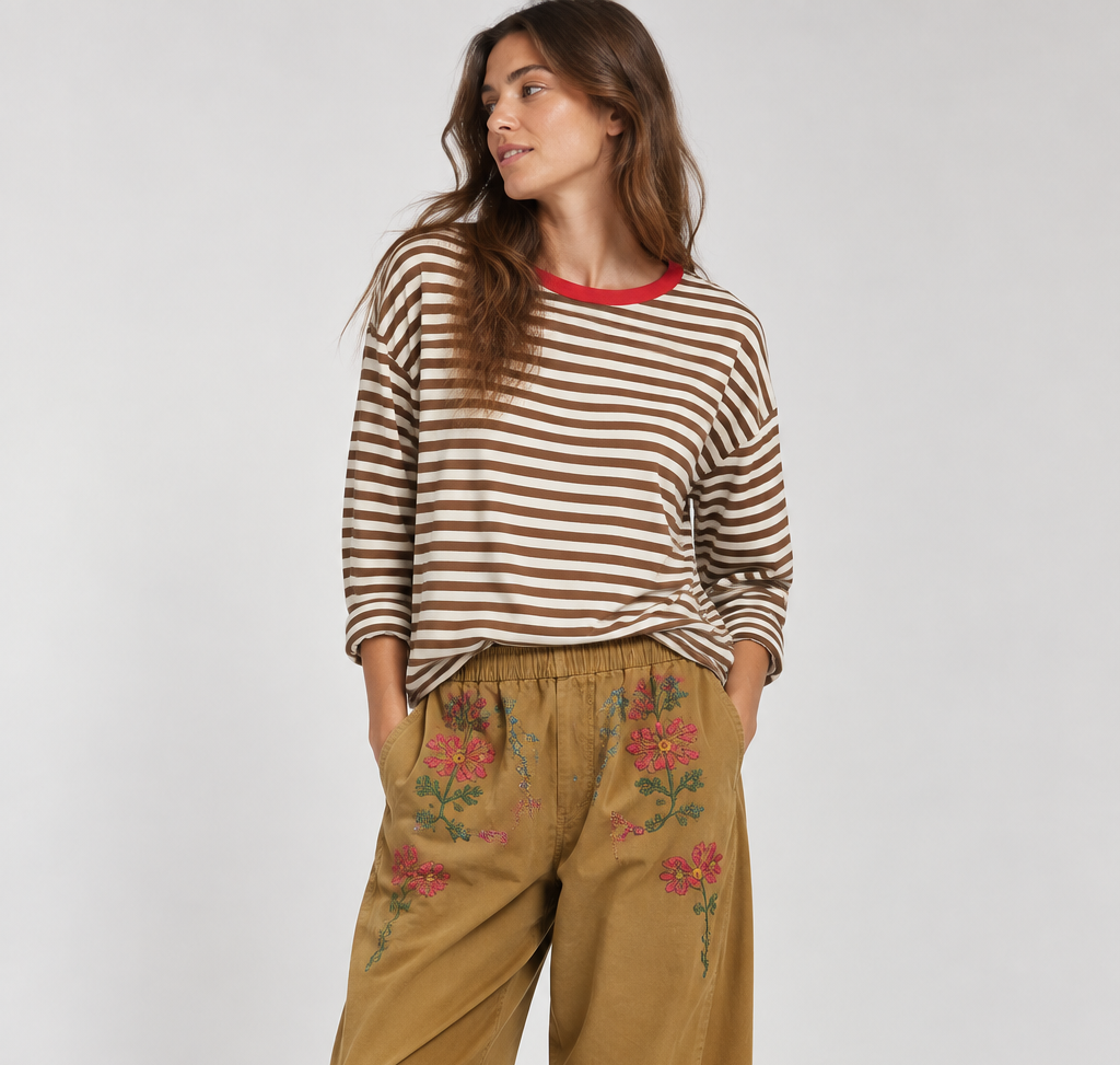 A woman modeling a brown and white striped long-sleeve top with a red collar, paired with wide-leg tan pants featuring floral embroidery on the thighs, and burgundy slip-on shoes, against a light gray studio background.