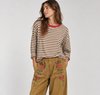A woman modeling a brown and white striped long-sleeve top with a red collar, paired with wide-leg tan pants featuring floral embroidery on the thighs, and burgundy slip-on shoes, against a light gray studio background.