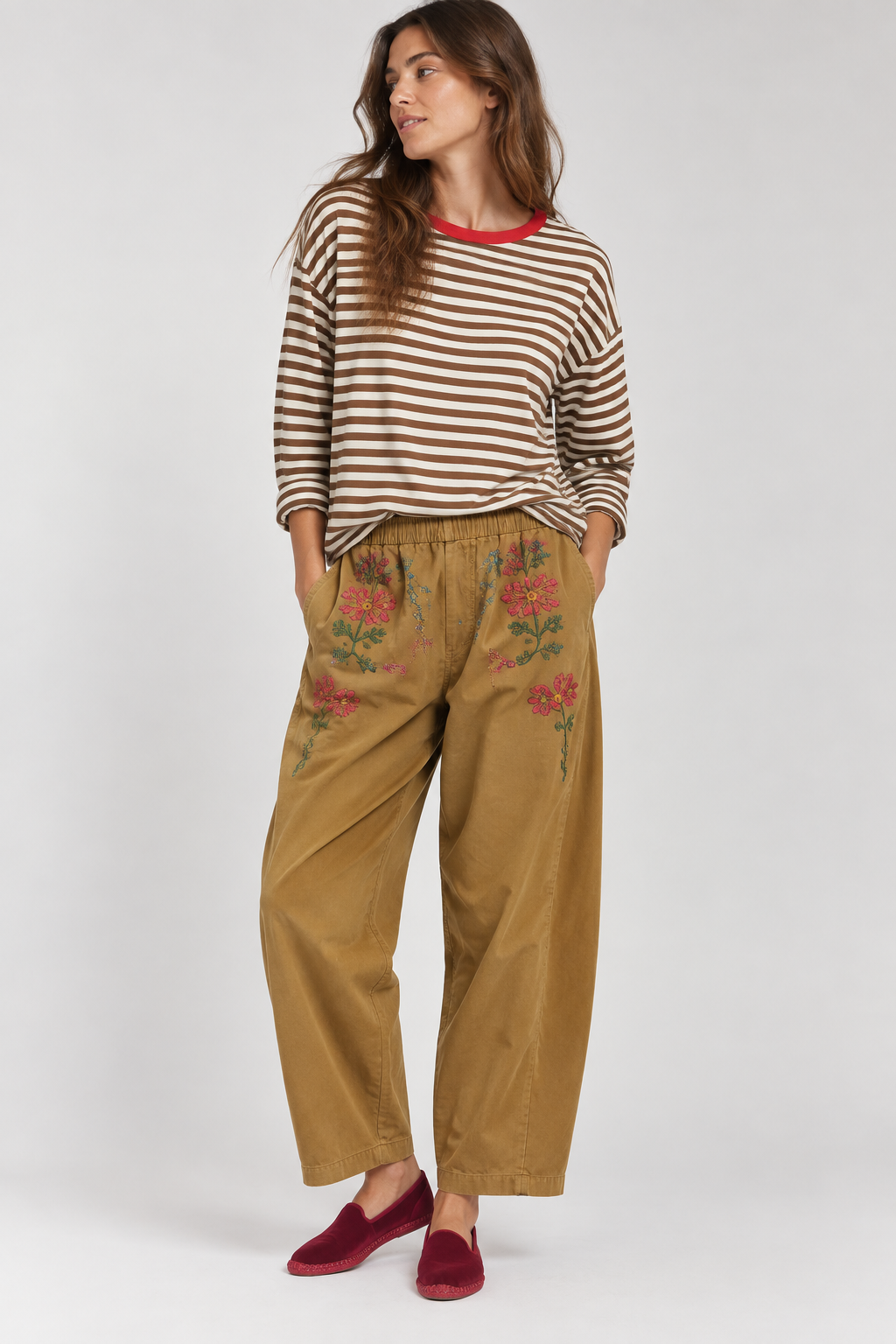 A woman modeling a brown and white striped long-sleeve top with a red collar, paired with wide-leg tan pants featuring floral embroidery on the thighs, and burgundy slip-on shoes, against a light gray studio background.