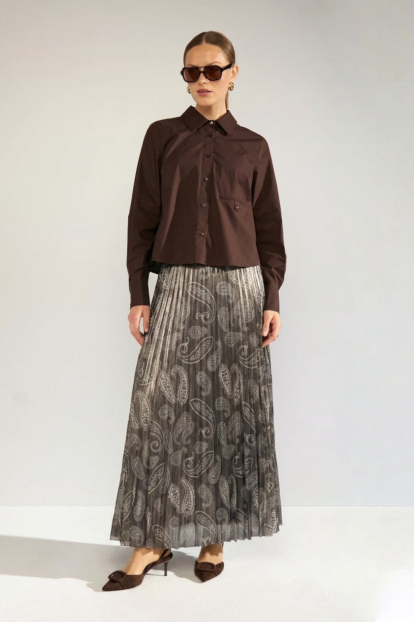 A woman modeling a dark brown long-sleeve button-up shirt paired with a pleated gray skirt featuring a paisley pattern, set against a plain light gray studio background.