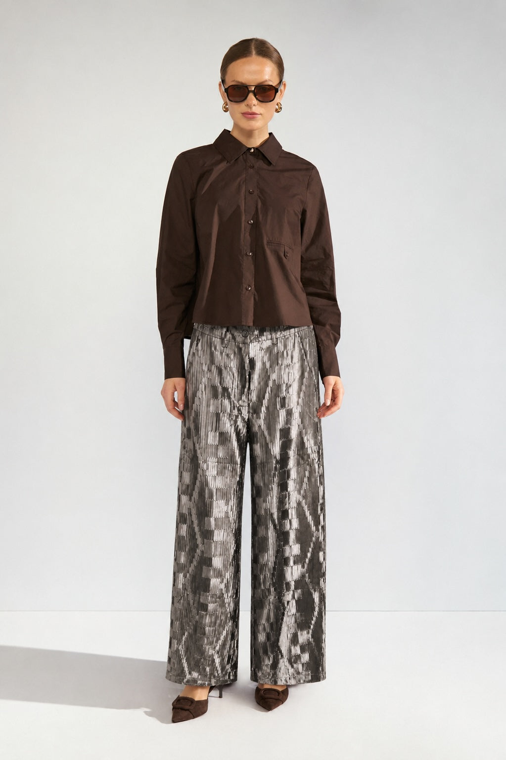 A model stands against a light gray studio background wearing a dark brown long-sleeve button-up shirt and wide-leg trousers with a gray and black snakeskin pattern. She is also wearing dark sunglasses and brown pointed-toe heels.