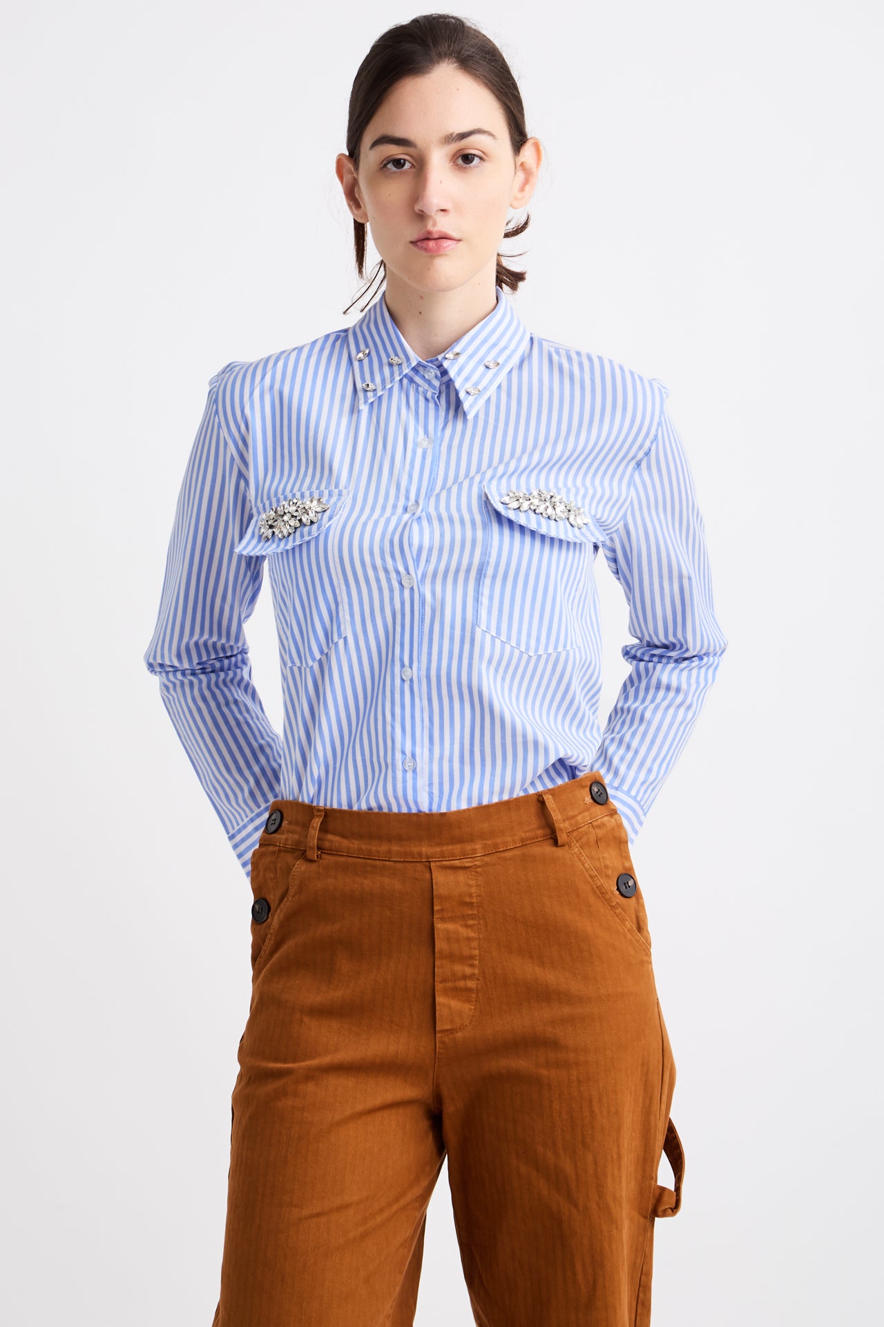 Person wearing a blue striped shirt with decorative elements and brown pants on a white background