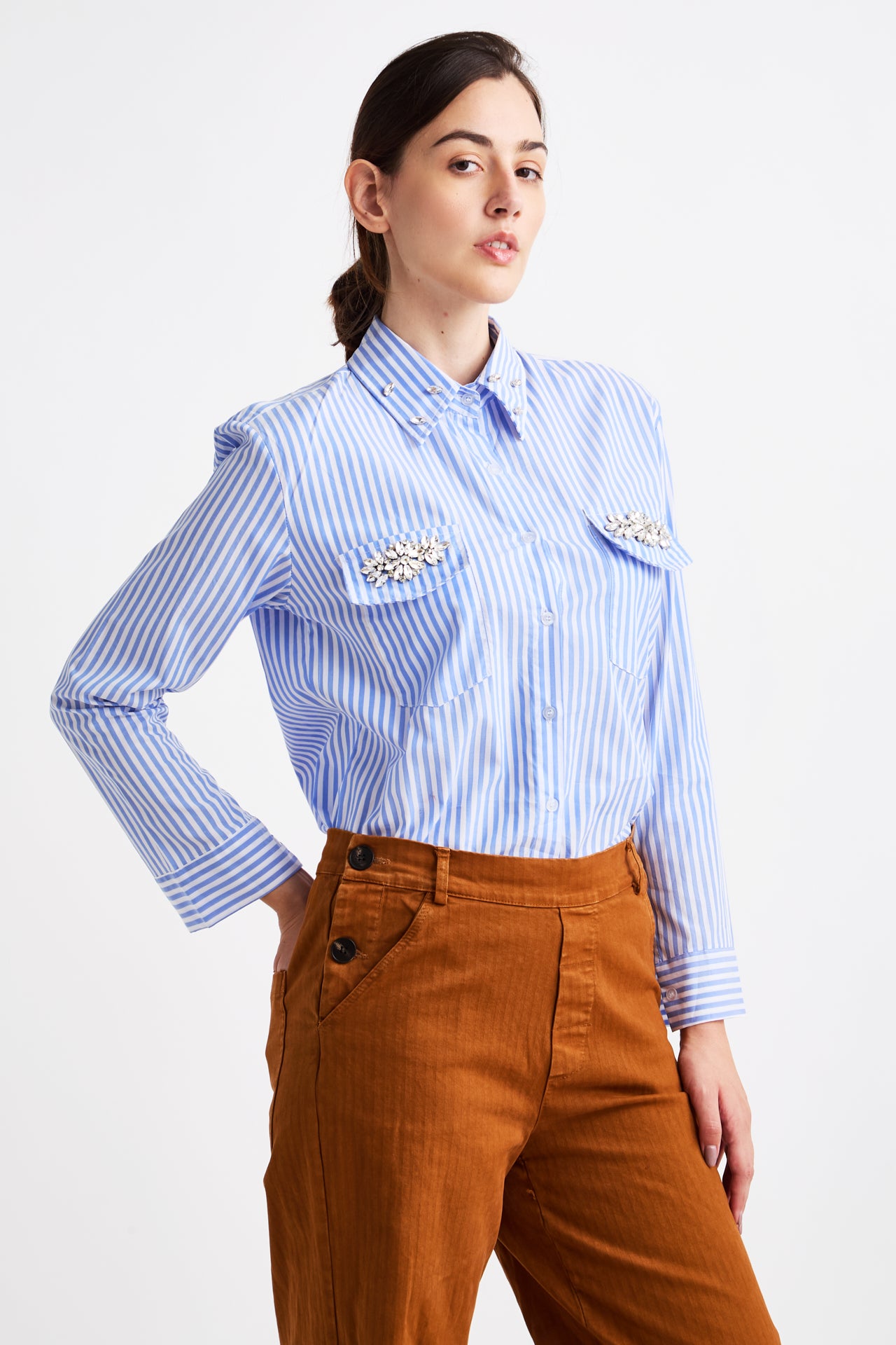 Woman wearing a blue and white striped shirt with decorative buttons and brown pants on a white background