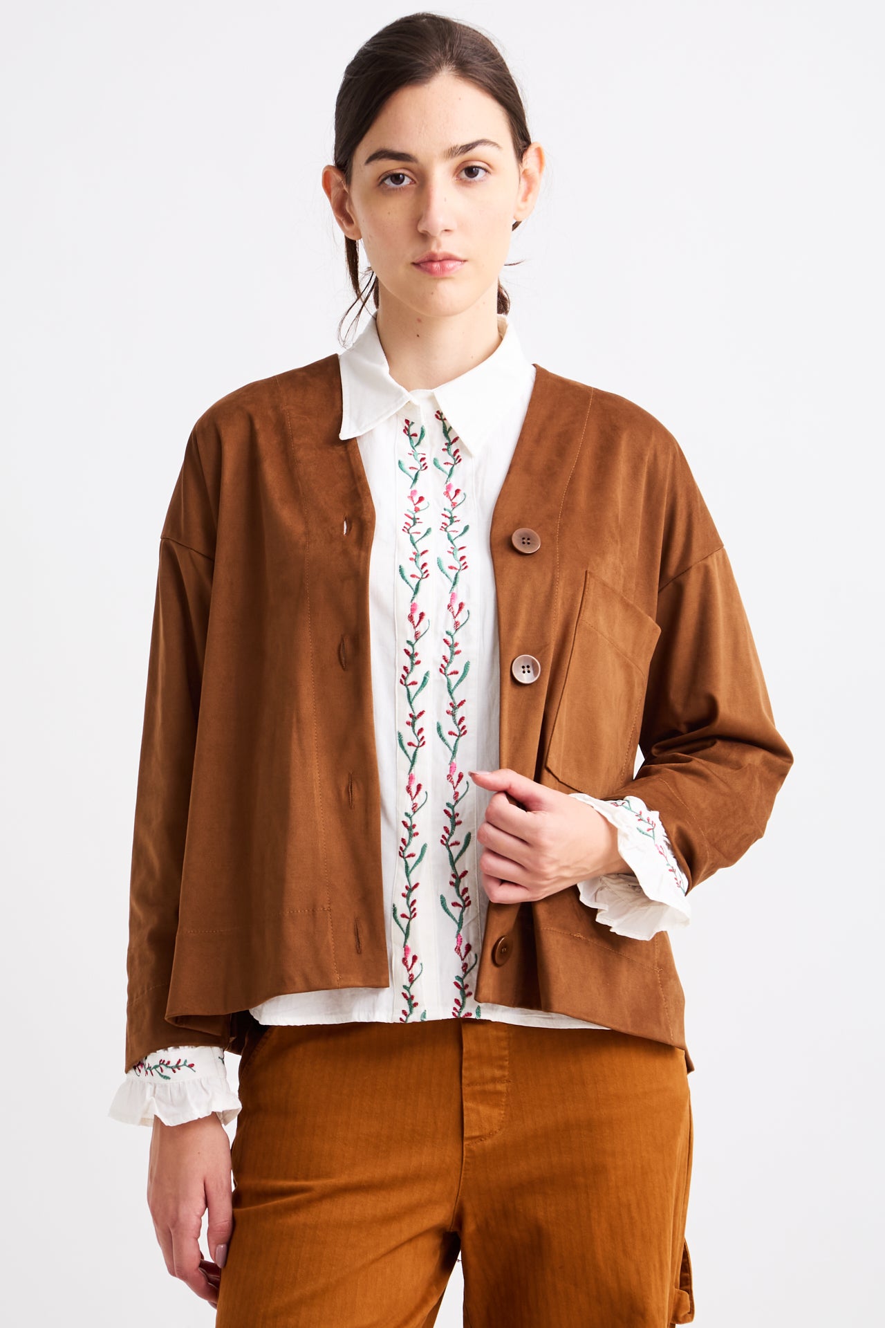 Person wearing a brown jacket over a white shirt with floral embroidery on a white background