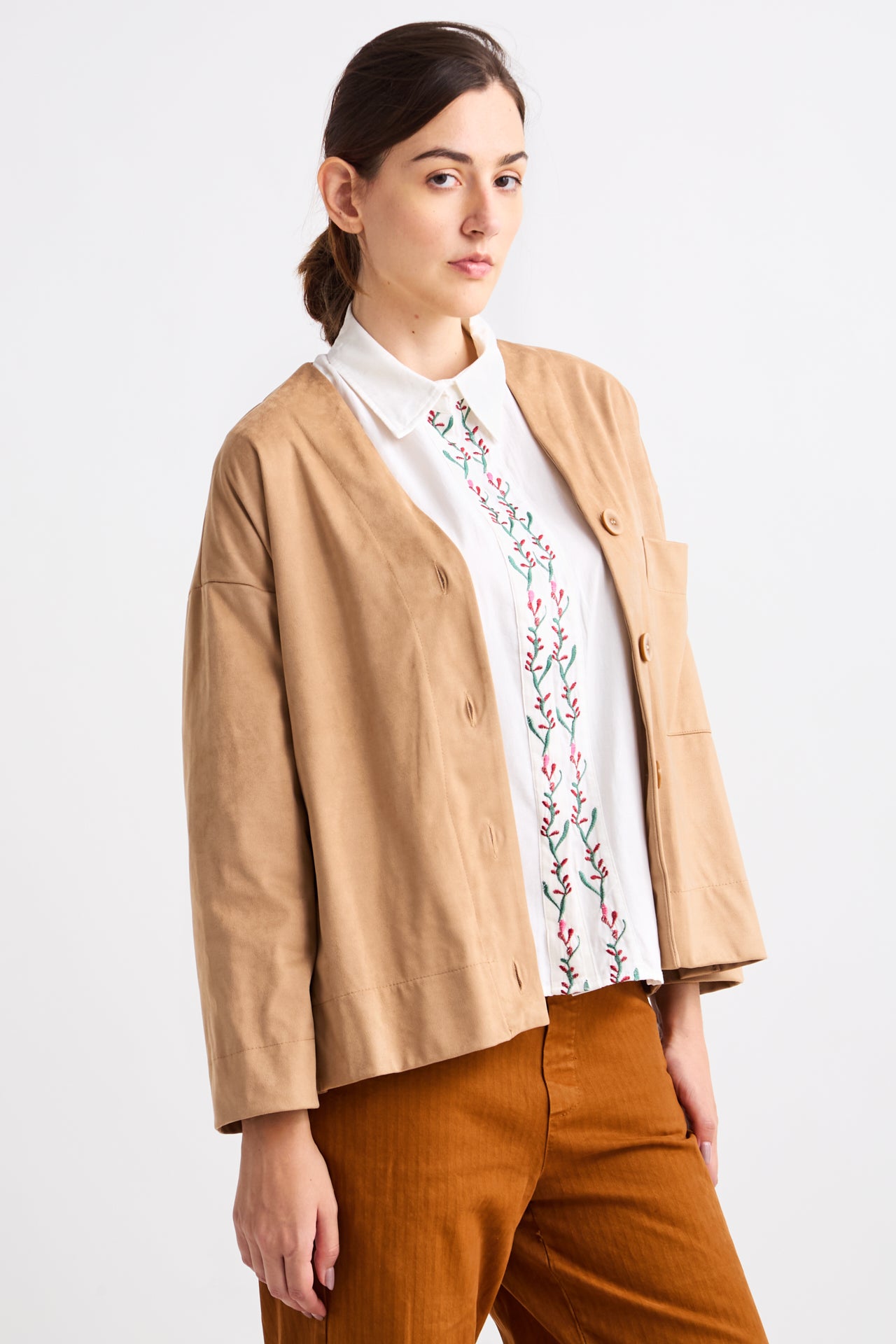 Woman wearing a beige jacket over a white shirt with floral embroidery on a white background