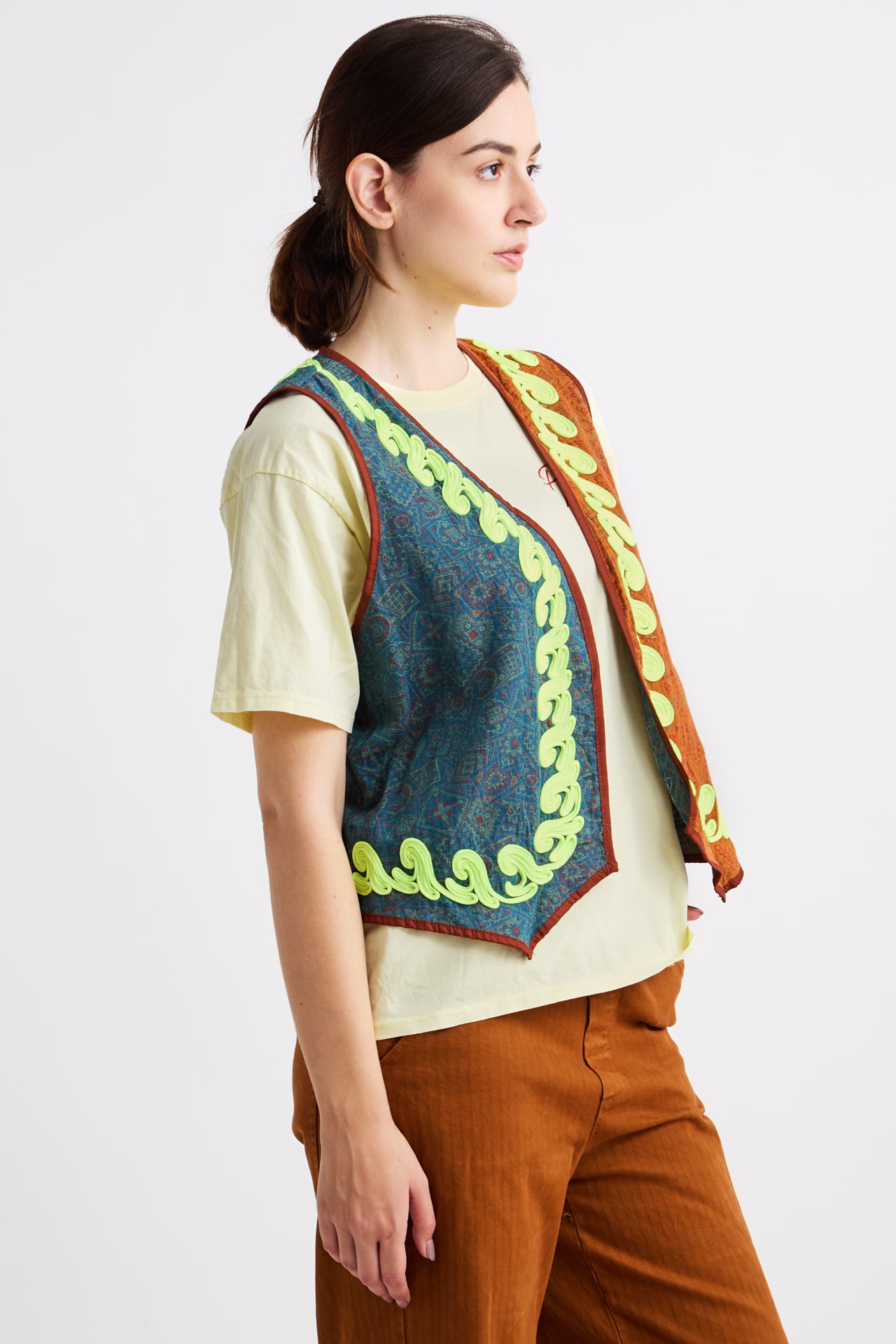 Woman wearing a colorful embroidered vest over a light shirt and brown pants on a white background