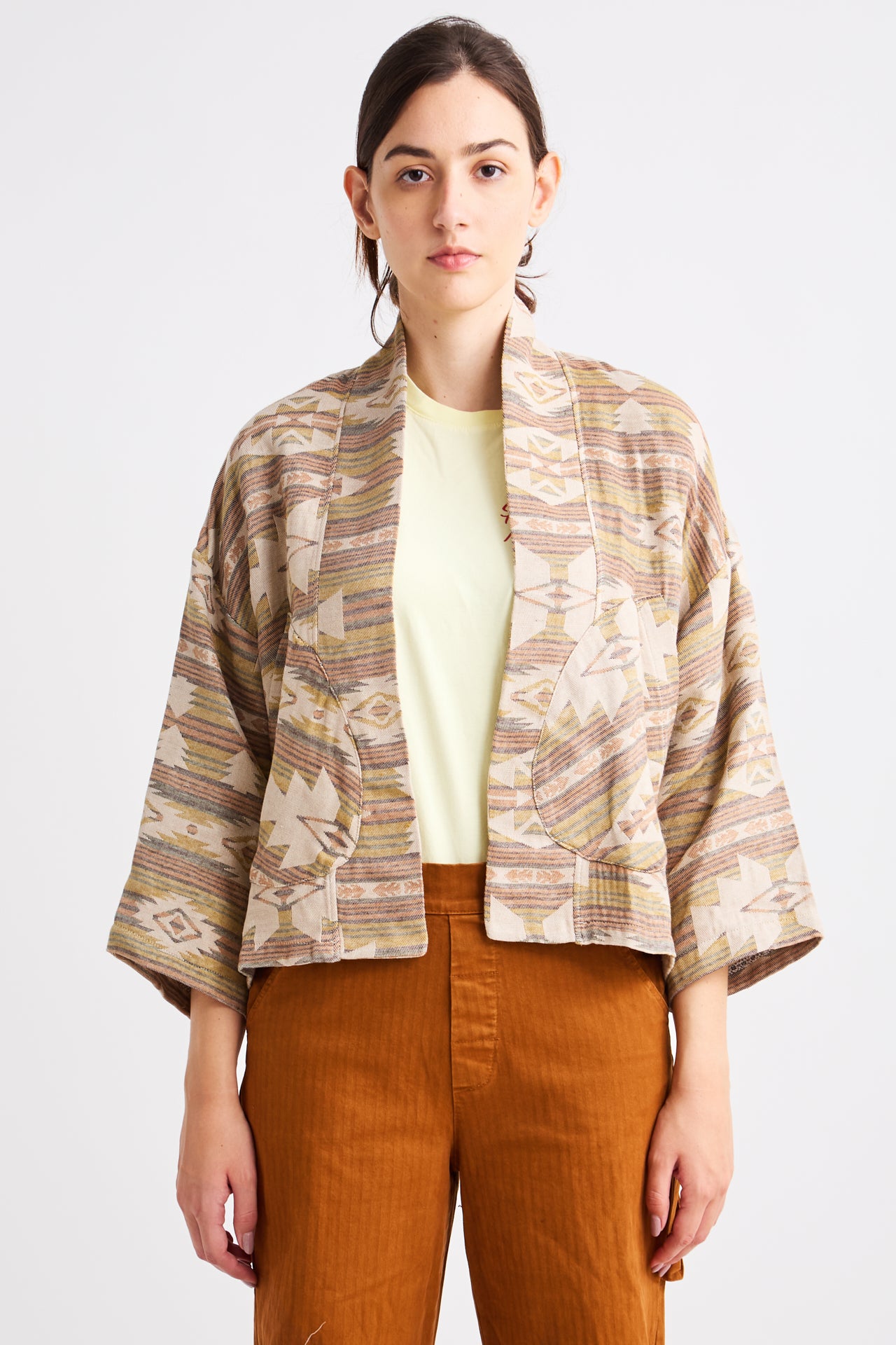 Woman wearing a patterned jacket over a yellow top and brown pants on a white background