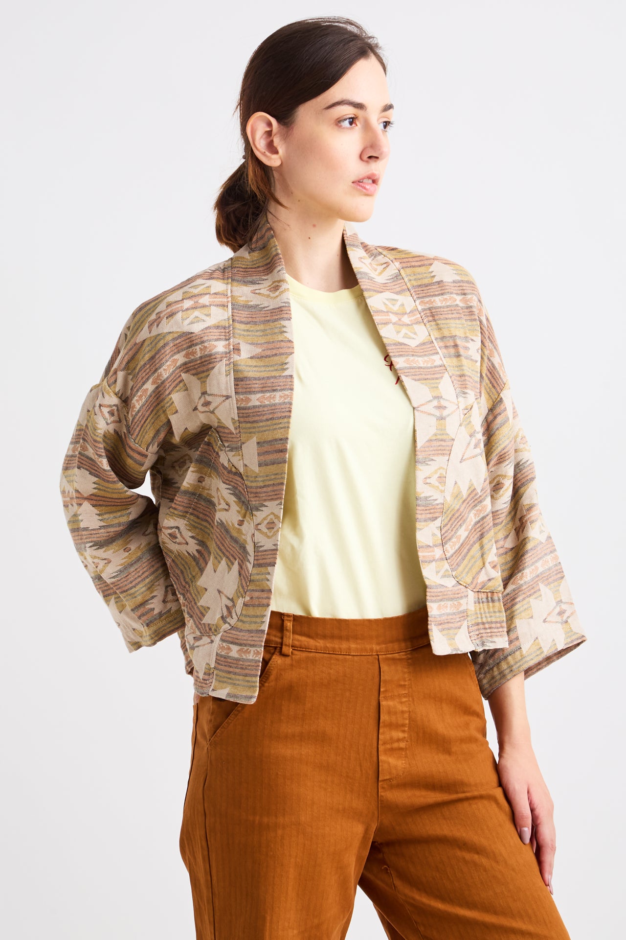 Woman wearing a patterned jacket over a yellow top and brown pants on a white background