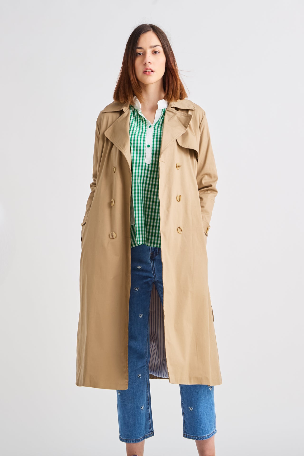 Woman wearing a beige trench coat over a green checkered shirt on a white background