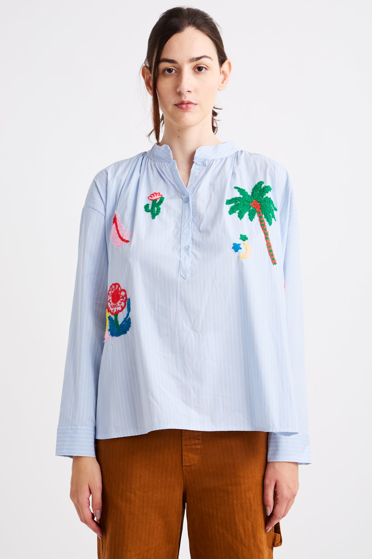 Woman wearing a light blue shirt with colorful floral and palm tree embroidery on a white background